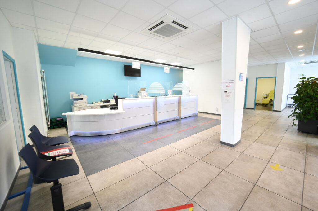 Espace de réception moderne et vide dans une clinique avec un bureau blanc, des murs bleus, un sol carrelé gris et des marqueurs de distanciation sociale au Laboratoire Reims Luton Bio Ard'Aisne.