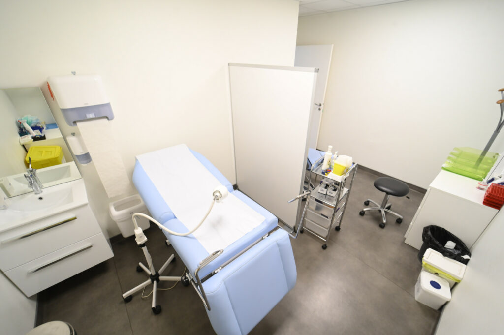 Une salle d'examen médical moderne avec un lit d'examen, des instruments médicaux sur plateau, un tabouret et des armoires au Laboratoire Reims Luton Bio Ard'Aisne.