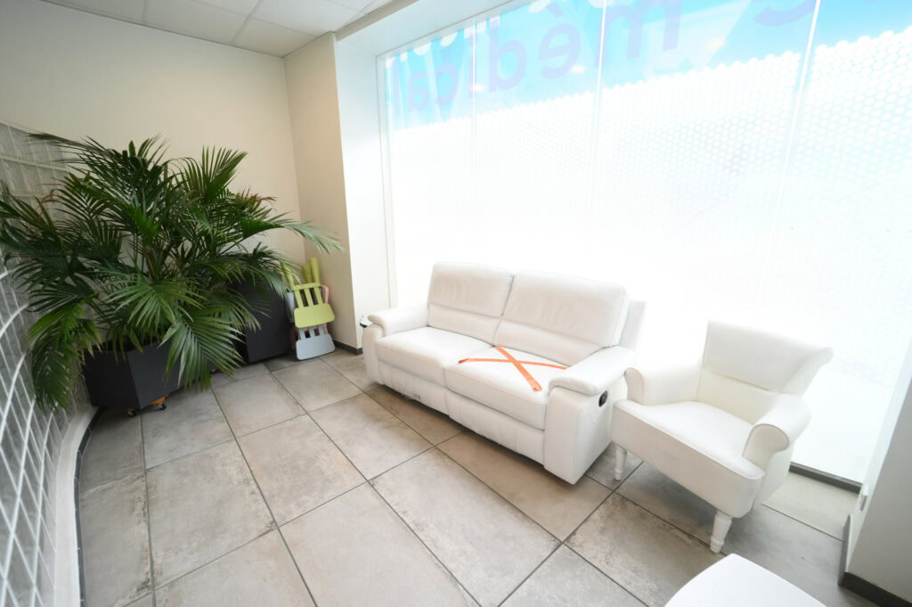 Un espace d'attente moderne avec un ensemble de canapés blancs, un cerceau, une grande plante en pot et des fenêtres vitrées avec une enseigne translucide du Laboratoire Reims Luton Bio Ard'Aisne.