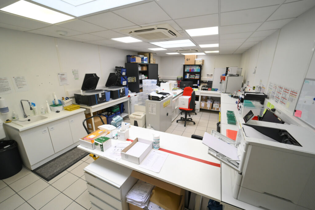 Une salle moderne du Laboratoire Reims Luton Bio Ard'Aisne, dotée d'ordinateurs, d'équipements médicaux et de zones de traitement des échantillons. Elle est organisée avec des tableaux sur les murs et une table de travail centrale.