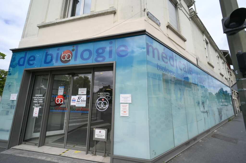 Vue extérieure d'un laboratoire de biologie médicale Reims Luton avec signalétique bleue, grandes baies vitrées affichant des images de la nature et portes d'entrée accessibles.