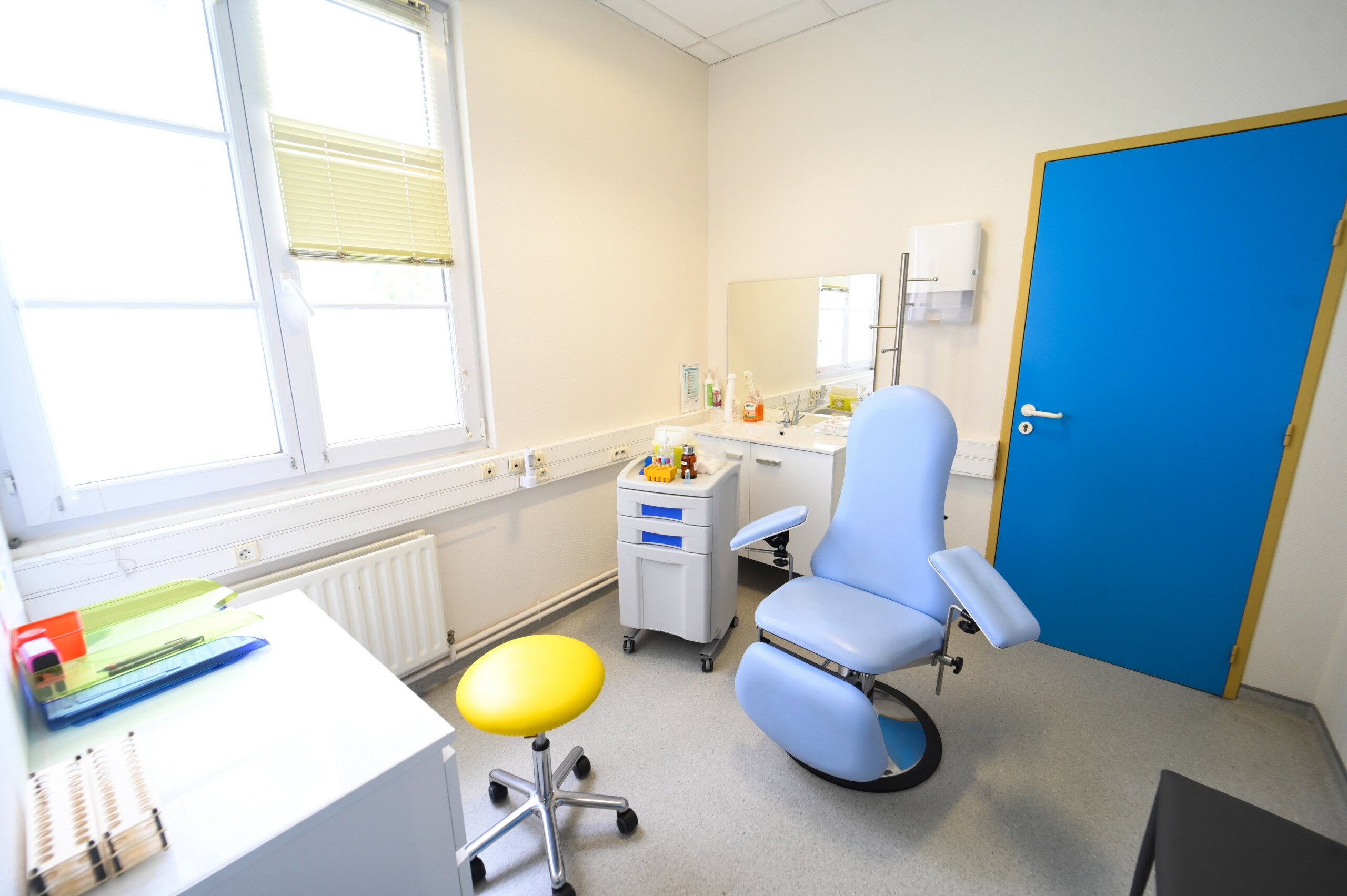Une salle d'examen médical lumineuse du Laboratoire Givet Bio Ard'Aisne avec une chaise bleue, un tabouret jaune, un bureau et des fournitures médicales près d'une fenêtre.