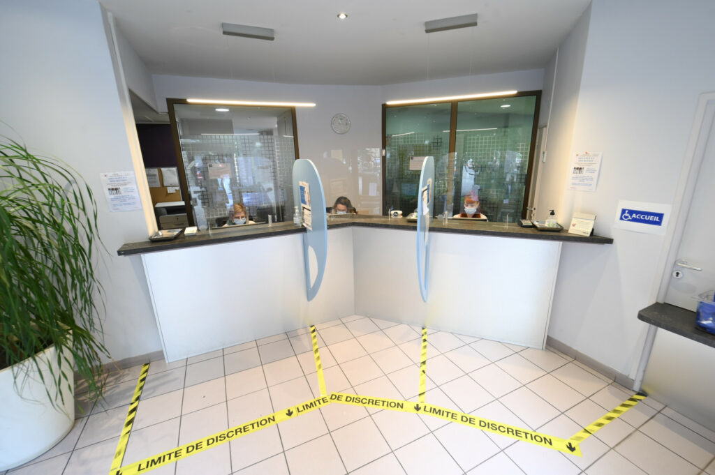 Vue intérieure d'un espace d'accueil avec cloisons vitrées, un préposé assis dans le Laboratoire Sedan La Marck et un marquage jaune au sol indiquant les limites de discrétion.