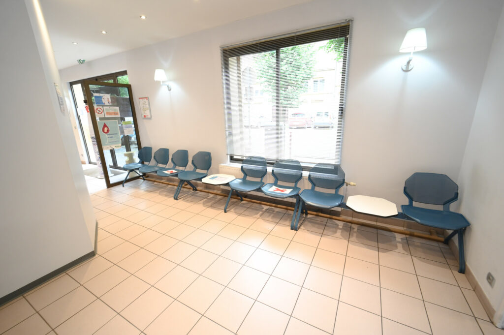 Salle d'attente lumineuse avec chaises bleues alignées contre un mur blanc, magazines sur une petite table, carrelage au sol et porte vitrée à l'entrée du Laboratoire Sedan La Marck.