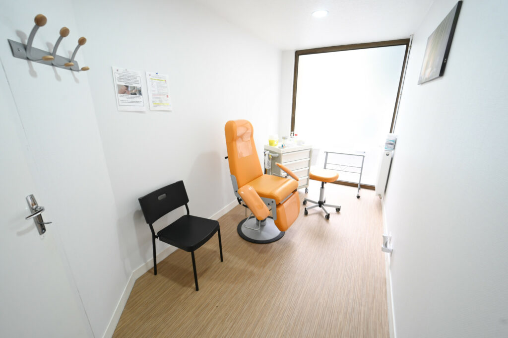 Une salle d'examen médical moderne avec une chaise réglable orange, une chaise noire, un bureau avec du matériel du Laboratoire Sedan La Marck Bio Ard'Aisne et des affiches d'information au mur.