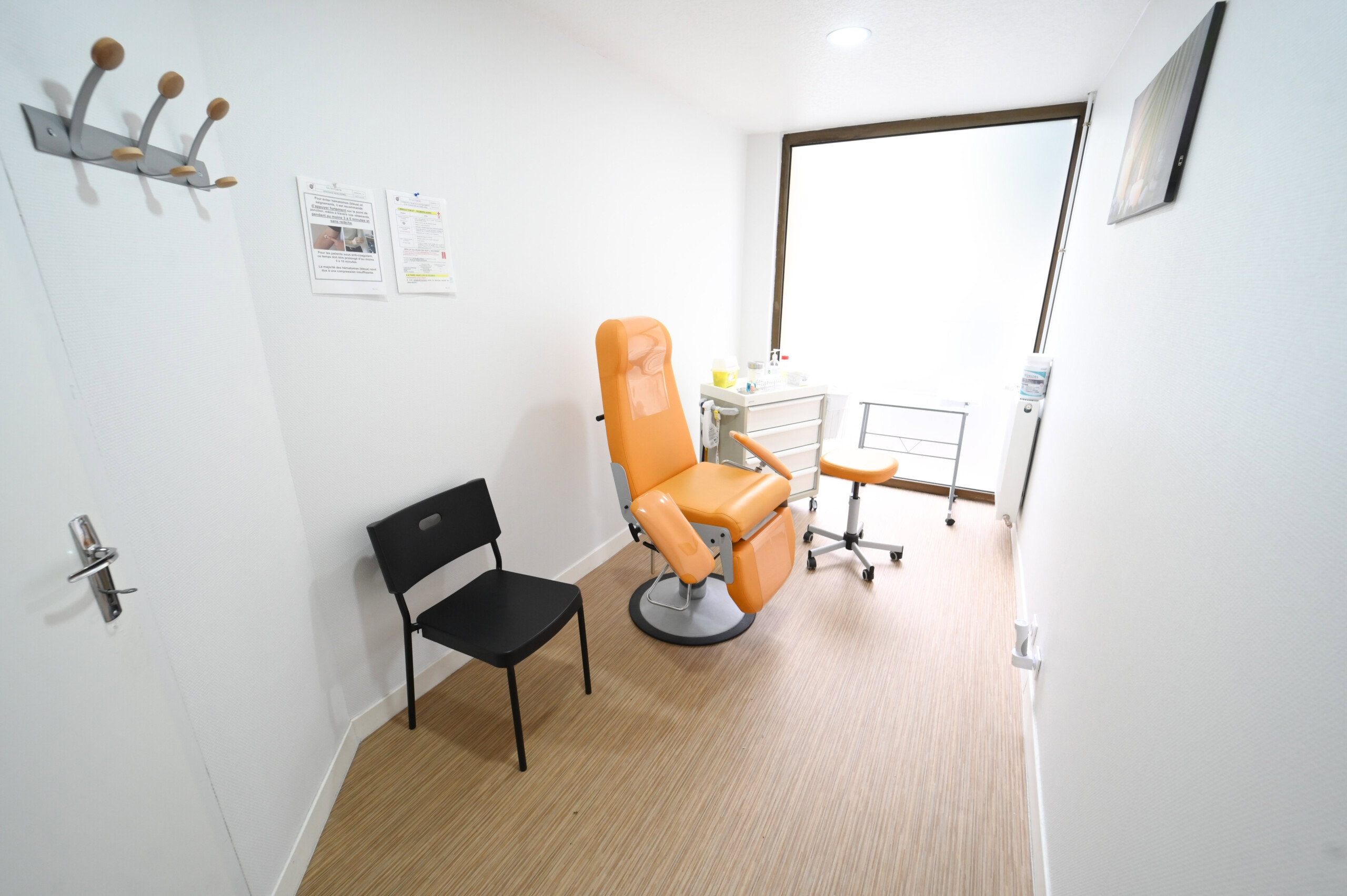Une salle d'examen médical moderne avec une chaise réglable orange, une chaise noire, un bureau avec du matériel du Laboratoire Sedan La Marck Bio Ard'Aisne et des affiches d'information au mur.