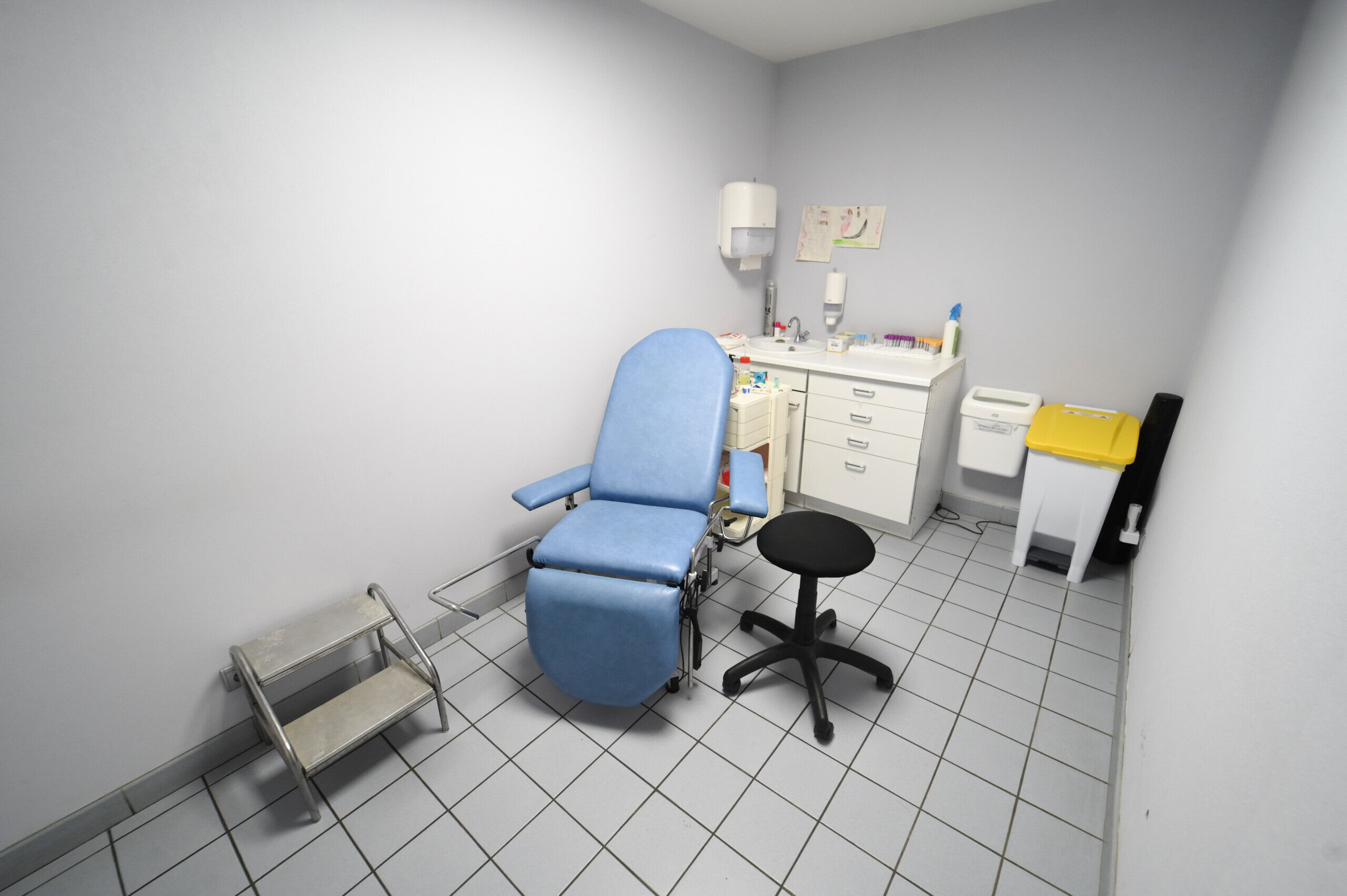 Une petite salle clinique avec un fauteuil d'examen bleu, un tabouret, des armoires blanches et des fournitures médicales sur un sol carrelé au Laboratoire Sedan La Marck.