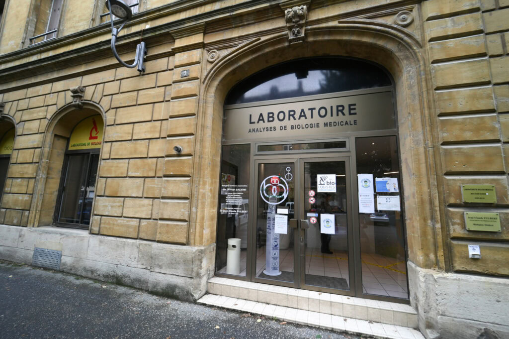 Vue de face de l'entrée du Laboratoire Sedan La Marck avec portes vitrées, plusieurs panneaux et façade en pierre sur une rue de la ville.