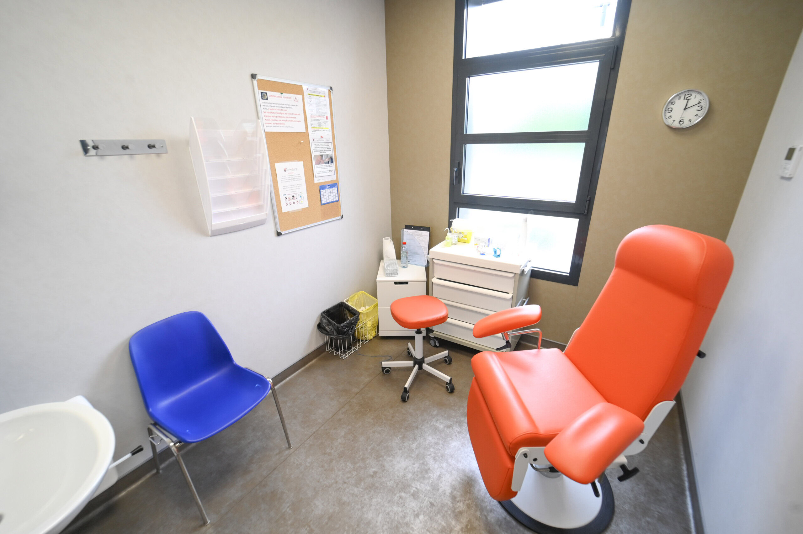 Une salle d'examen médical du Laboratoire Balan Bio Ard'Aisne comprend une chaise réglable rouge, une chaise latérale bleue, un petit bureau avec des fournitures médicales, une horloge au mur et un