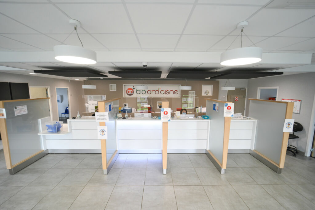 Un intérieur de pharmacie moderne avec un comptoir blanc, des écrans de protection et une signalétique favorisant la santé et le bien-être au Laboratoire Balan Bio Ard'Aisne.