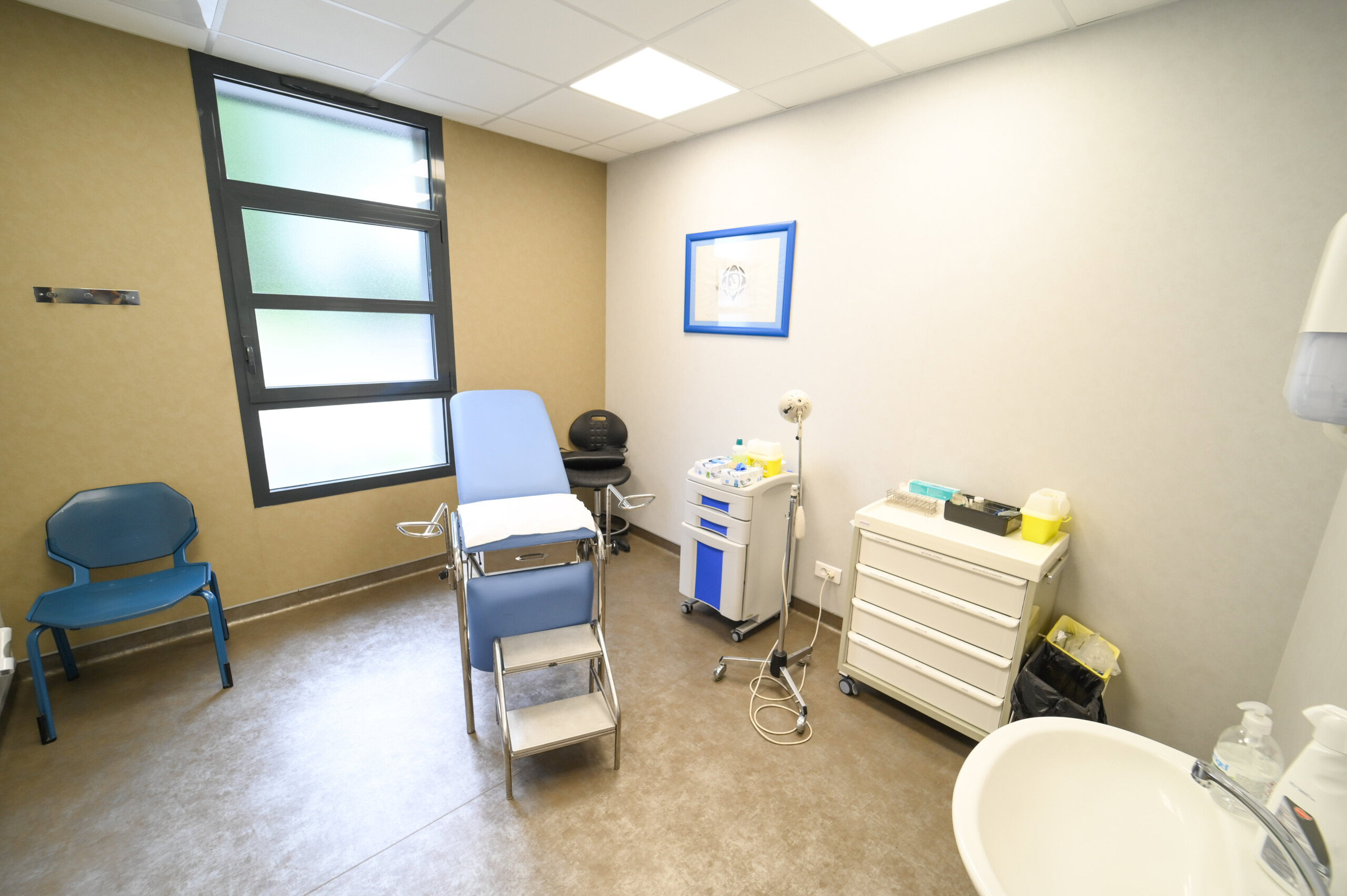 Une salle d'examen médical moderne avec une chaise de patient, des instruments médicaux sur un chariot, un évier et deux chaises bleues à côté d'une grande fenêtre au Laboratoire Balan Bio Ard'Aisne.