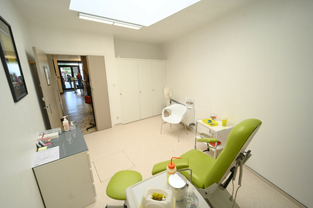 Une salle médicale lumineuse avec un fauteuil d'examen vert, des armoires blanches et du matériel médical du Laboratoire Vouziers Bio Ard'Aisne, menant à une porte ouverte.