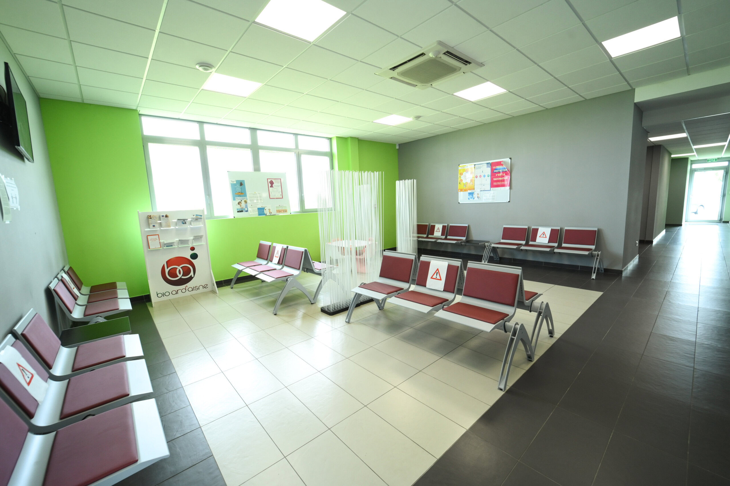 Salle d'attente bien éclairée avec murs verts, plusieurs rangées de chaises rouges et argentées et bibliothèques avec les magazines du Laboratoire Rethel Bio Ard'Aisne.