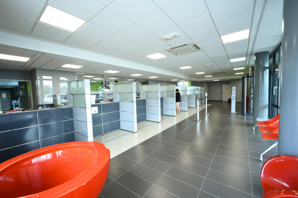 Hall d'entrée moderne du Laboratoire Rethel Bio Ard'Aisne avec des cabines bleues, des chaises orange le long du mur et une personne debout à un comptoir éloigné.