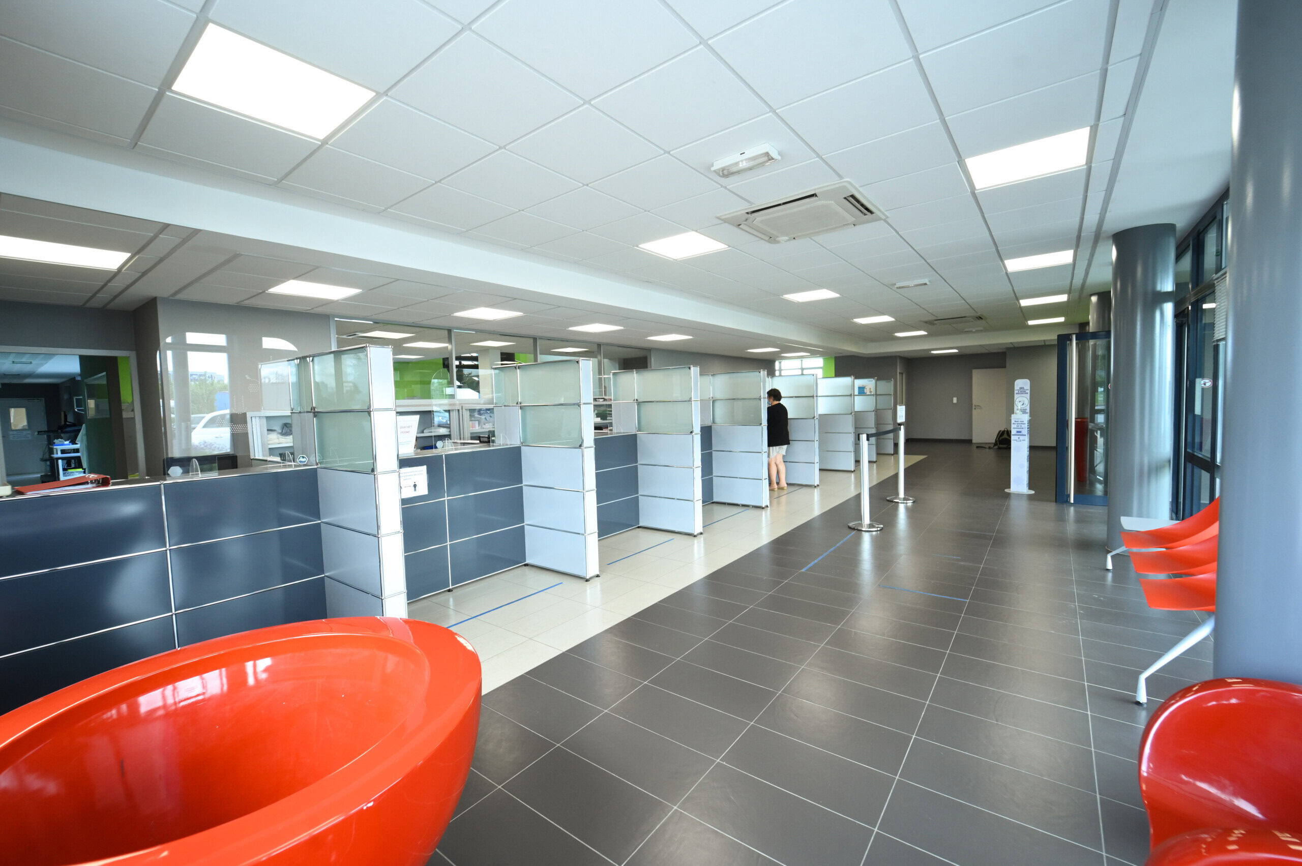 Hall d'entrée moderne du Laboratoire Rethel Bio Ard'Aisne avec des cabines bleues, des chaises orange le long du mur et une personne debout à un comptoir éloigné.