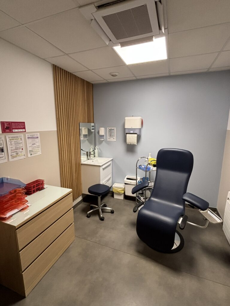 Une Salle D’examen Médical Avec Une Chaise, Un Tabouret, Un Lavabo Et Diverses Fournitures Médicales Sur Des étagères Et Des Comptoirs.