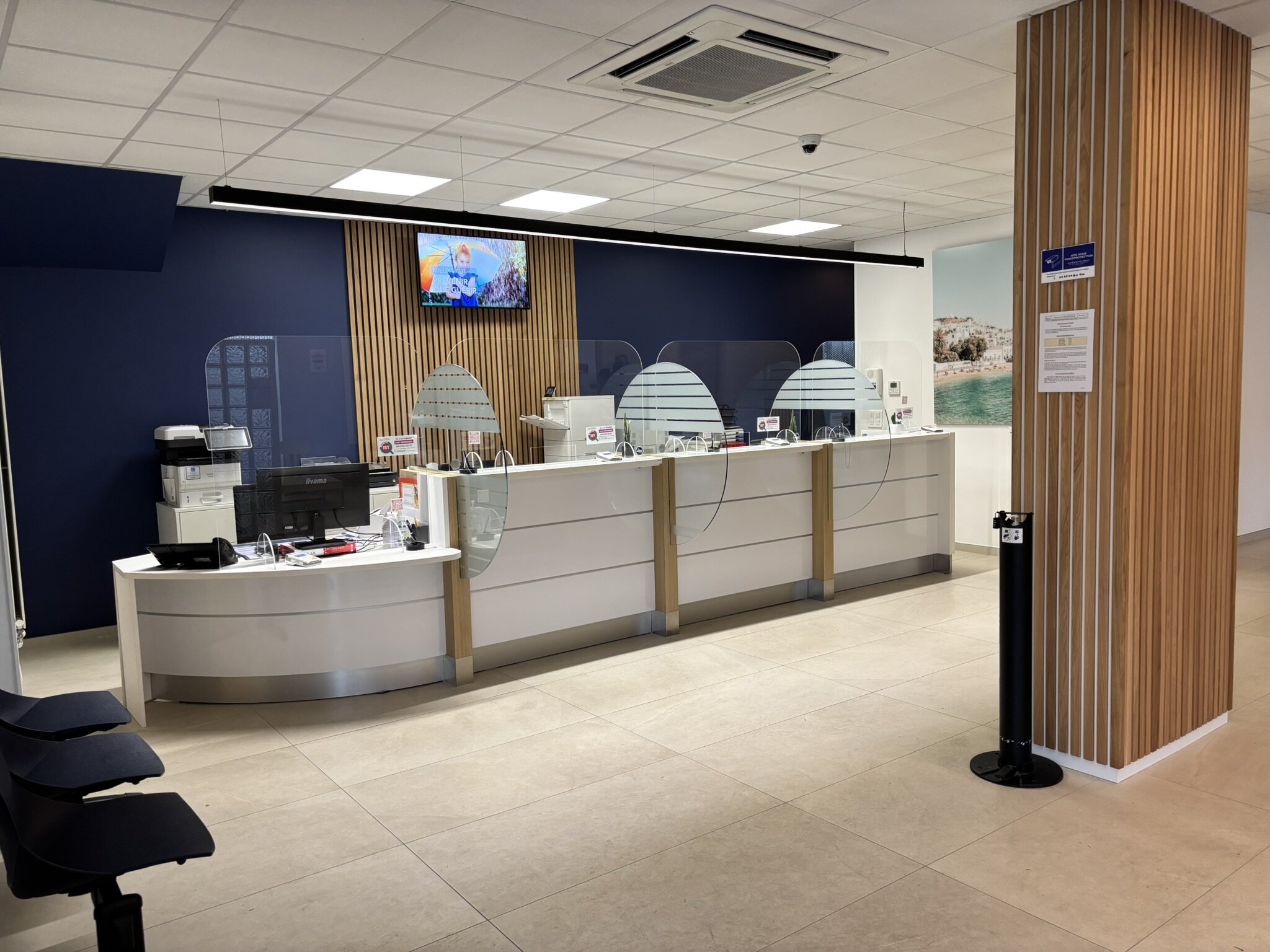 Un Intérieur De Banque Moderne Avec Un Comptoir De Réception, Des Barrières De Protection En Verre, Un écran De Télévision Sur Un Mur Bleu Foncé Et Un Sol En Carrelage Beige. Trois Chaises Noires Font Face Au Comptoir.