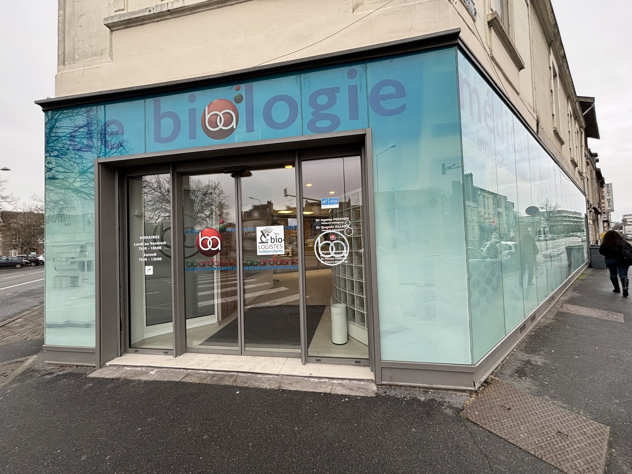 Vue De La Rue D'un Immeuble D'angle Moderne Aux Murs En Verre, Avec Le Mot « Biologie » Et Divers Logos Sur La Façade. Piéton Marchant à Proximité Par Temps Nuageux.