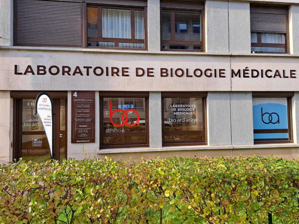 Vue extérieure d'un laboratoire de biologie médicale avec une grande enseigne, un logo et une porte d'entrée, derrière une haie verte devant le bâtiment.